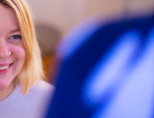 Das halbe, grinsende Gesicht einer Mitarbeiterin vor einem unscharfen Monitor